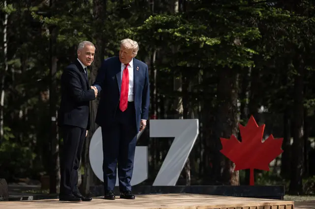 Prime Minister Mark Carney shakes hands with Donald Trump at the G7 in Alberta. They are both wearing suits. A white "G7" sign and a red maple leaf sign is behind them.