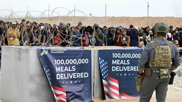 A crowd of Palestinians stands near a wall, armed by a man in military uniform. Signs are strung up reading: '100,000,000 meals delivered'