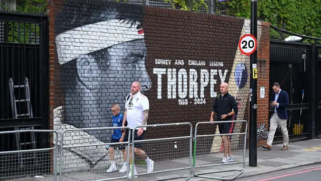 Fans walk past a Graham Thorpe mural outside the Oval. It reads 'Surrey and England legend Thorpey 1969-2024' with artwork of him in black and white on the left hand side.