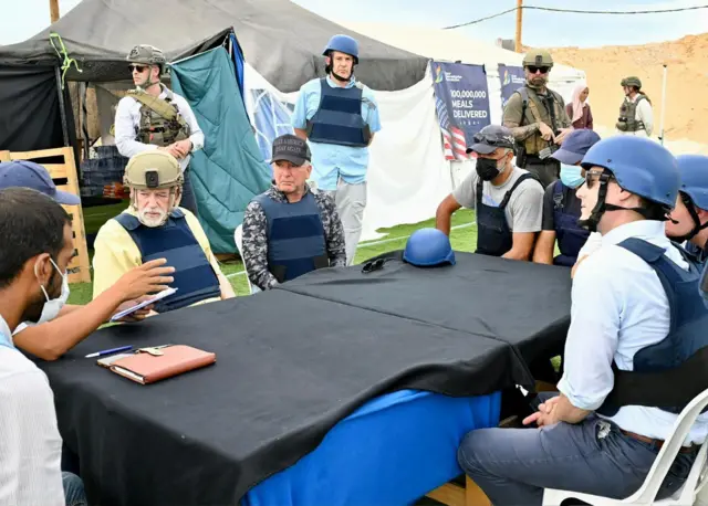 Steve Witkoff sites US Ambassador to Israel Mike Huckabee at a table at an aid distribution site, along with several ther men. They are all wearing bodyarmour and Witkoff is wearing a camo top and baseball cap.