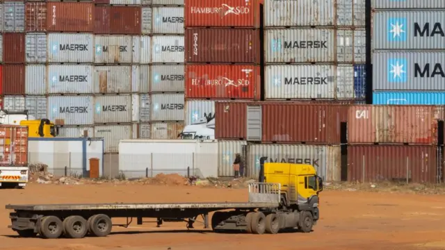 rows of shipping containers can be seen. a flat bed truck drives in front of them.
