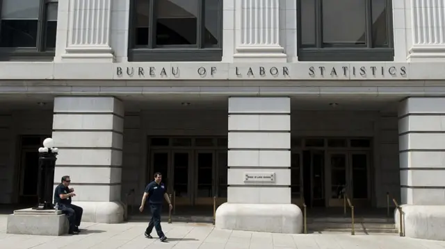 Bureau of Labor Statistics building, which is written on the it in black lettering. The building is light grey stone. There are two officials outside it, one on the left sitting down and another on the centre-left walking away from the building