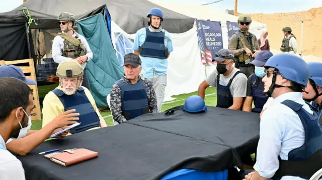 Witkoff and Huckabee sitting at a makeshift table covered in a black cloth, with six other people also sitting around it. Two are wearing helmets, three in caps, and three in face masks. There are four people standing. This is outside and behind them is a GHF tent