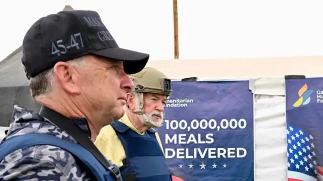 Witkoff and Huckabee on the left, facing right. Witkoff in the forground wearing a black MAGA cap, black and grey military camouflage shirt and a bulletproof vest. Huckabee is wearing a khaki coloured helmet, a pastel yellow shirt and a bulletproof vest. behind them is a GHF poster that reads 100,000,000 meals delivered.