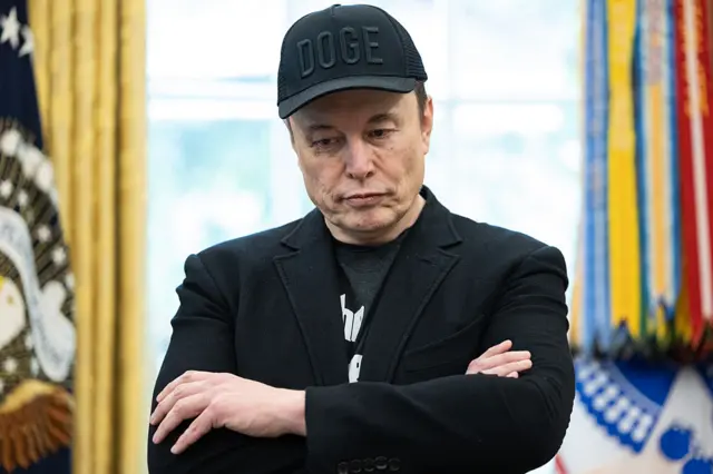 Elon Musk stands in the Oval Office, He is wearing a baseball cap with the word "DOGE" written on it and is wearing a dark coat.