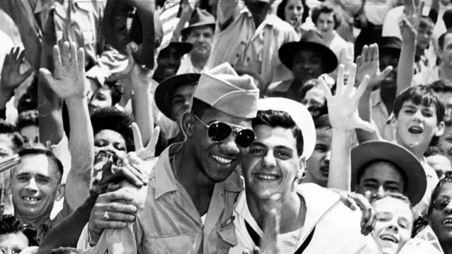 Soldiers cheer and wave towards the camera. An black man in uniform with aviator sunglasses (L) with his right arm around a smiling white sailor (R)