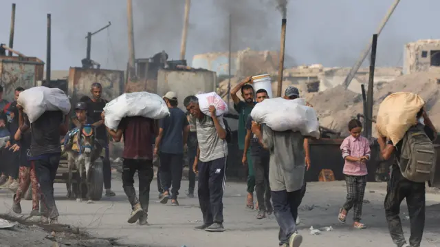 Men carry white parcels of aid on their shoulders. Some children can also be seen. Rubble is in the background with dark smoke rising
