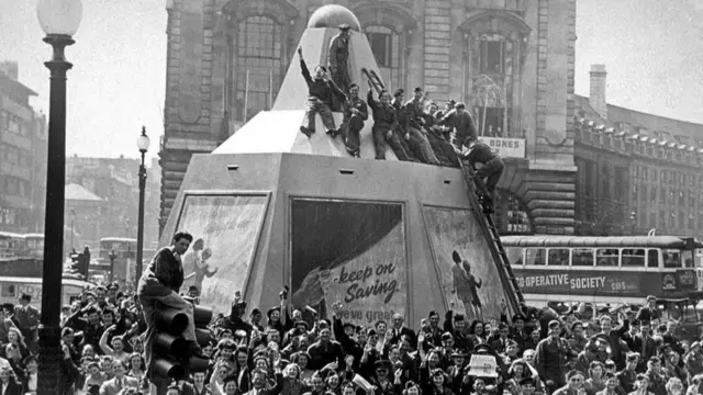 A large group celebrating at Piccadilly Circus. Some soldiers can be seen sitting on top of a wooden carcass protecting the Shaftesbury Memorial Fountain. There’s posters attached to the carcass, including one reading “Keep Saving”