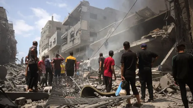 A group of men wearing t-shirts and shorts stood facing away from camera. In front of them is a damaged building with smoke rising. Scattered around them is debris from the building including bricks and metal.