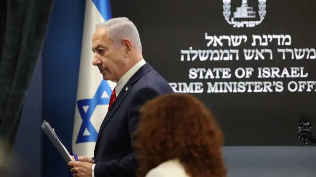 Netanyahu exits a press briefing. He is holding papers.