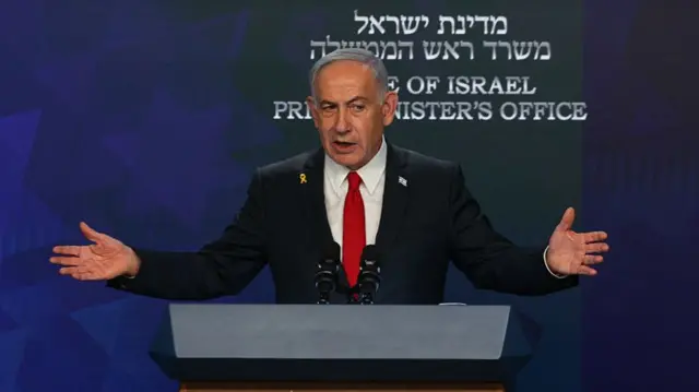 Benjamin Netanyahu stood behind a podium, arms outstretched wearing a dark suit with a white shirt and red tie. Behind him is a label written in Hebrew and below in English that reads Israel Prime Minister's Office.
