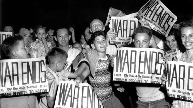 A group of children, women and men look to the camera as they hold copies of The Knoxville Journal with ‘War Ends’ written on them