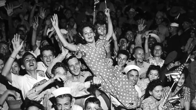 A crowd look towards the camera with their hands in the air as they celebrate. A woman in a polka dot dress (C) is hoisted over the shoulders of a sailor, another woman smiling holding her from the armpits