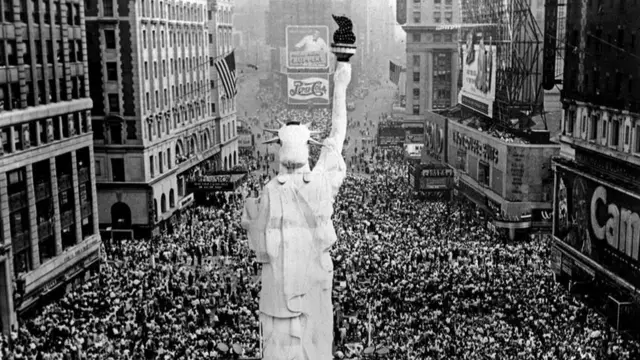 A shot of the scaled-down version of the Statue of Liberty from behind with a huge crowd below it, Times Square visible in the background, Broadway to the left and 7th Avenue to the right