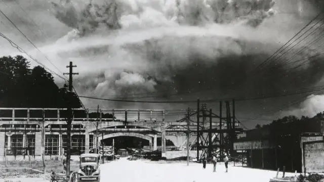 A black and white photo shows buildings, power lines and a car with large black clouds in the sky above