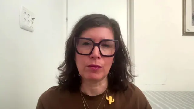 Sharone Lifschitz looks directly at the camera with a white wall and door behind her. She has long frizzy hair and big black spectacles