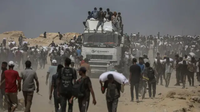 Crowds of Palestinians walk by a white aid truck, many carrying white sacks of aid on their shoulders. A number of people sit atop the truck