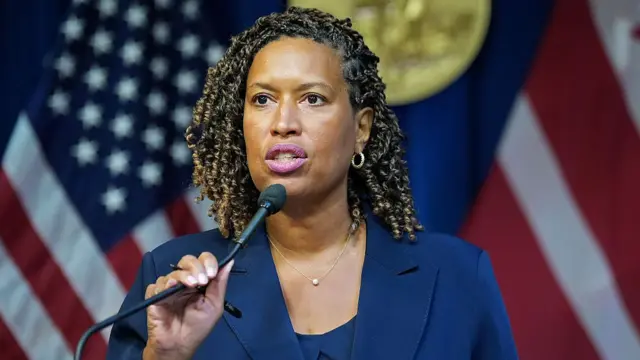 DC mayor Muriel Bowser in dark blue blazer and matching shirt speaks into a thin black mic, her right hand positioning it in front of her. In the blurred background are two blurred US flags