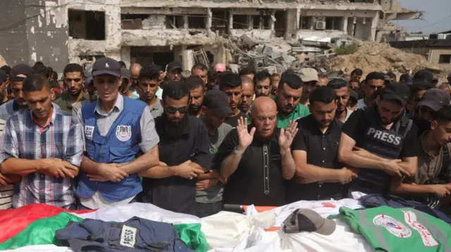 A crowd of people stand by the shrouds containing the bodies of the journalists. A press vest lies across one, and those crowded around look mournful