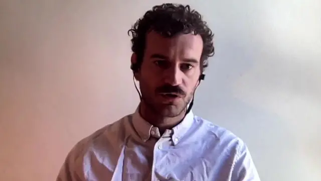 Roux, a man with curly brown hair and waring a white button-up shirt, speaks to the camera