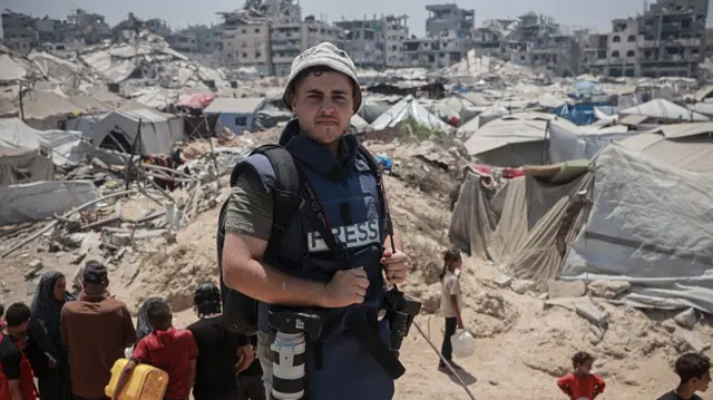 A man in a blue press bullet proof vest standing on a hill holding multiple cameras. There are people behind him looking over a huge dirty patch of land covered in tents. In the background are rows of destroyed buildings.