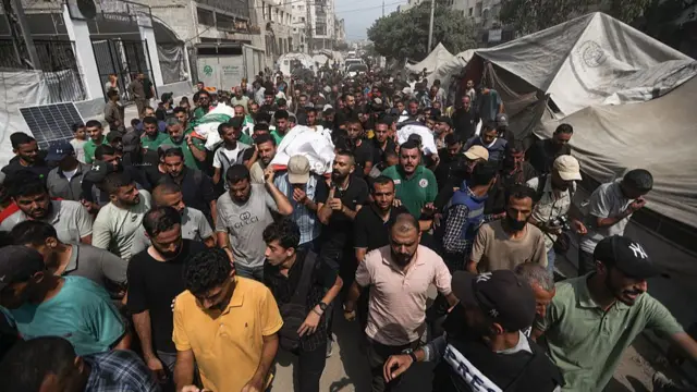 A crowd of people escort those carrying three white shrouds containing some of the bodies of the journalists through Gaza, past damaged buildings and tattered tents