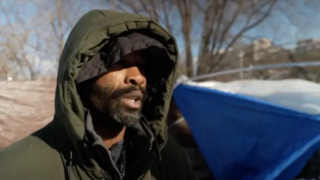 A man with a beard wears two coats in front of a snow-covered tent