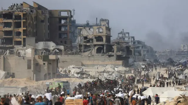 A scene looking down a dirt road that runs alongside a row of buildings. The road is full of people walking along in a dense crowd. There are tents lining the road as dust whips up around them. The buildings are totally destroyed, vacant with holes in them and crumbling down. In the distance around more buildings is smoke.