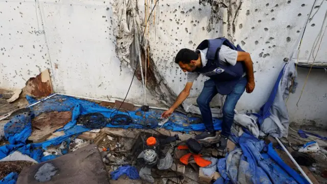 A man in a press vest stands in a bombed tent with rubble lying all over the flaw. He bends over, inspecting the mess. The concrete wall behind him is covered in holes that look like they were from bullets or shrapnel.