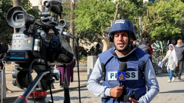 Mohammed Qreiqeh speaking into a microphone with Al Jazeera's logo on it, standing in front of a video camera. He is wearing a protective vest and a Hemet marked "presse"