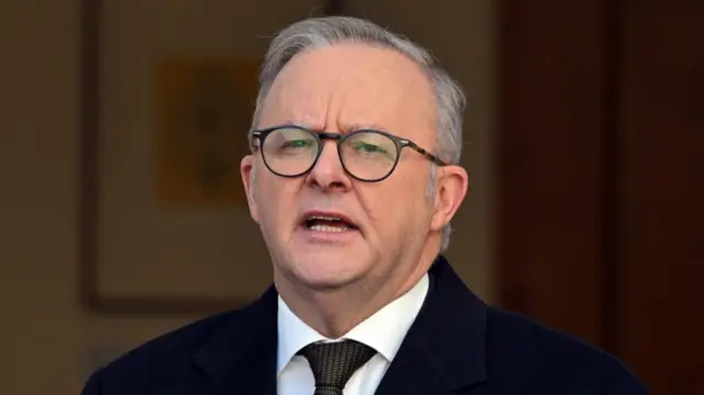 Headshot of Albanese speaking. He wears glasses and has grey hair. The background is blurry but it looks like there a picture on a wall.
