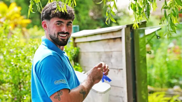 Frazer Walsh in a blue Chester Zoo branded polo shirt, holding food to feed otters. He has brown hair and is smiling at the camera, in front of a glass enclosure, surrounded by trees.