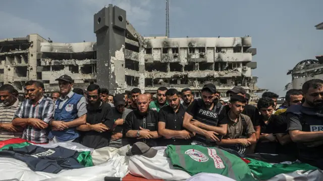 Mourners stand in front of the bodies of killed people, wrapped in white shrouds. They have their arms crossed, two are wearing press vests. The bodies have hats, a press vest and an emergency worker uniform on them. In the background is an almost entirely destroyed building.