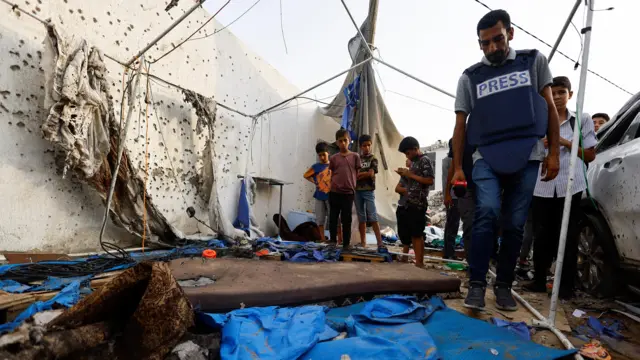 A man in a blue press vest looks at debris on the floor including blue tent material, some children look on from the background