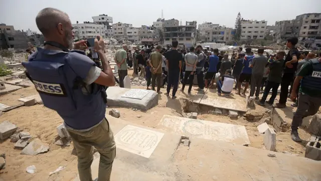 A man in a blue press vests stands a little away from a crowd, pointing a camera at them as they gather around a sandy grave