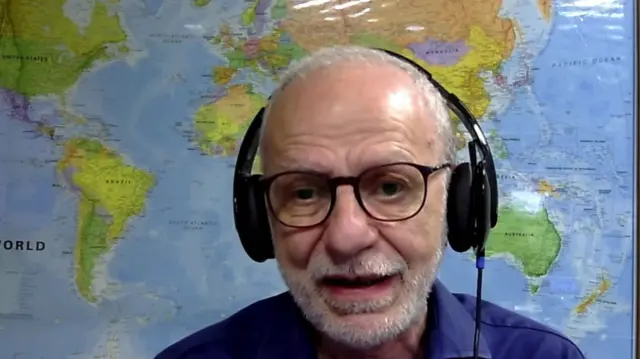 Headshot of Negm who sits with big black headphones on, in front of a picture of a world map