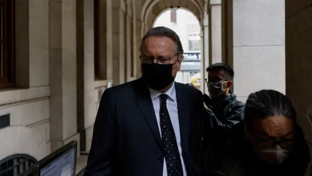 Timothy Owen wearing a black mask and dark suit and tie. He is looking to the side while photographers stand near him.