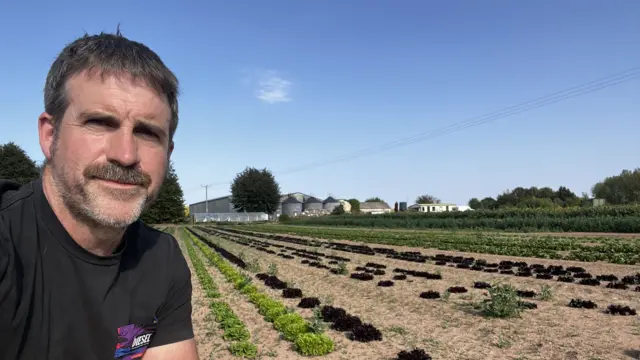 A man in a black t-shirt with a moustache takes a selfie next to rows of planted vegetables