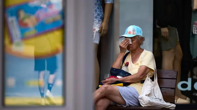 A woman wipes her face amid a heatwave, while she is sat on a wooden bench and wearing a blue peaked cap