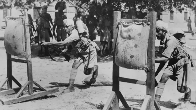 Black and white image of two Indian soldiers sticking sandbags during bayonet practice while other soldiers observe