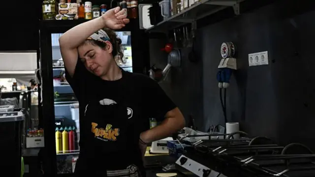 Woman stands in a professional kitchen, with her head raised against her head to wipe sweat from her forehead