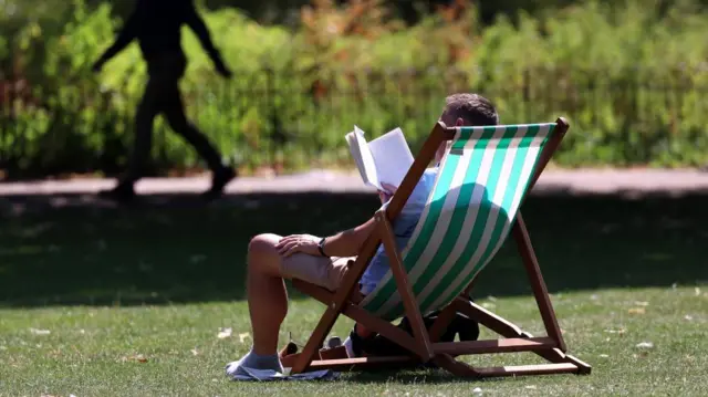 A man reclines in a sun chair in a park, reading a book