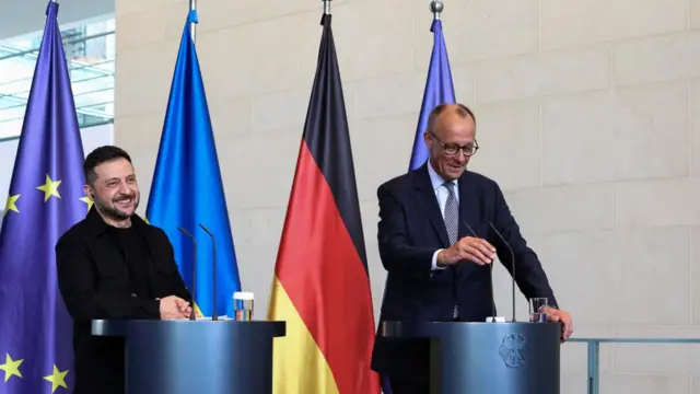 Zelensky and Merz both smiling as they speak into mics at two podiums. The flags of the EU, Germany and Ukraine are behind them as is a plain brick wall