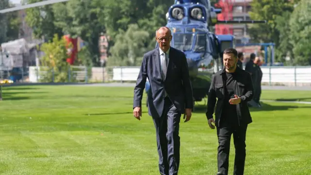 Zelensky and Merz talk as they walk along grass in front of a helicopter behind them