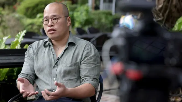 Sebastien Lai with round spectacles, in a green button up shirt, gesturing with his hands while sitting outdoors. In the background are some plants.
