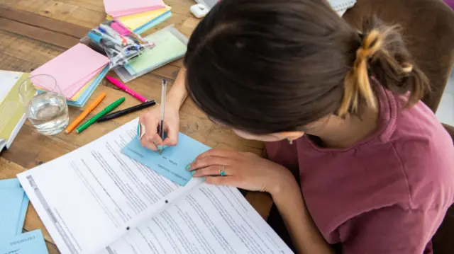 A girl sits at a table studying. she is filling out cue cards and looking over a review book