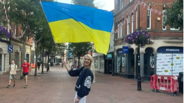 A blonde woman waves a large Ukrainian flag on a pedestrianised street