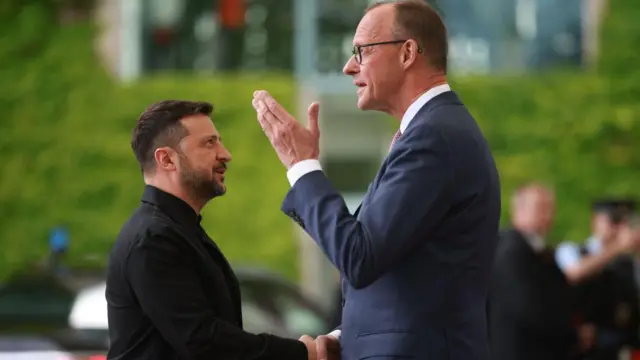 German Chancellor Friedrich Merz (R) welcomes Ukrainian President Volodymyr Zelensky. Zelensky is dressed in black and looking up at Merz, and shaking his hand while Merz uses his other hand to gesture.