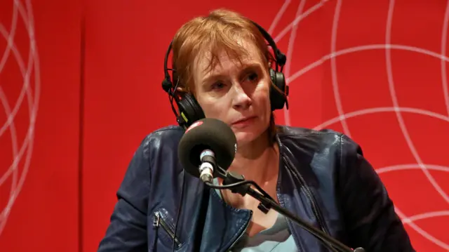 A woman in a navy blue jacket in front of a red background sits a microphone - she has short red hair