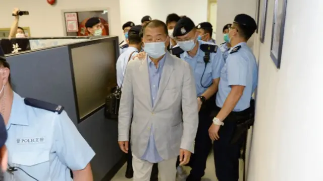 Jimmy Lai wearing a grey suit and face mask, surrounded by policemen in blue uniform along a corridor of an office.